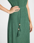 A close-up of a woman wearing a green pleated maxi dress with a tie-waist belt and tassel detail, shown against a plain light background.