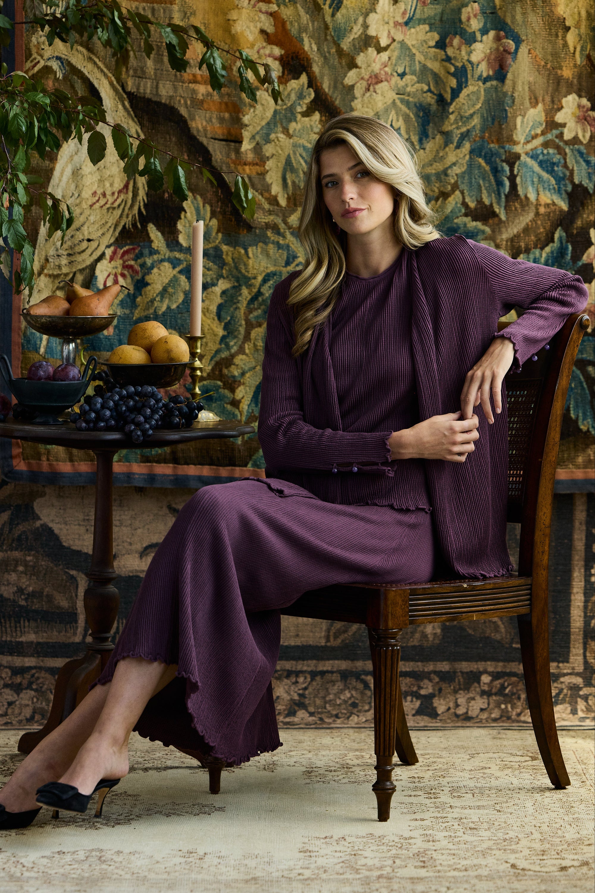 Woman sitting in a wooden chair against a patterned background wearing a pleated cardigan, skirt, and shell