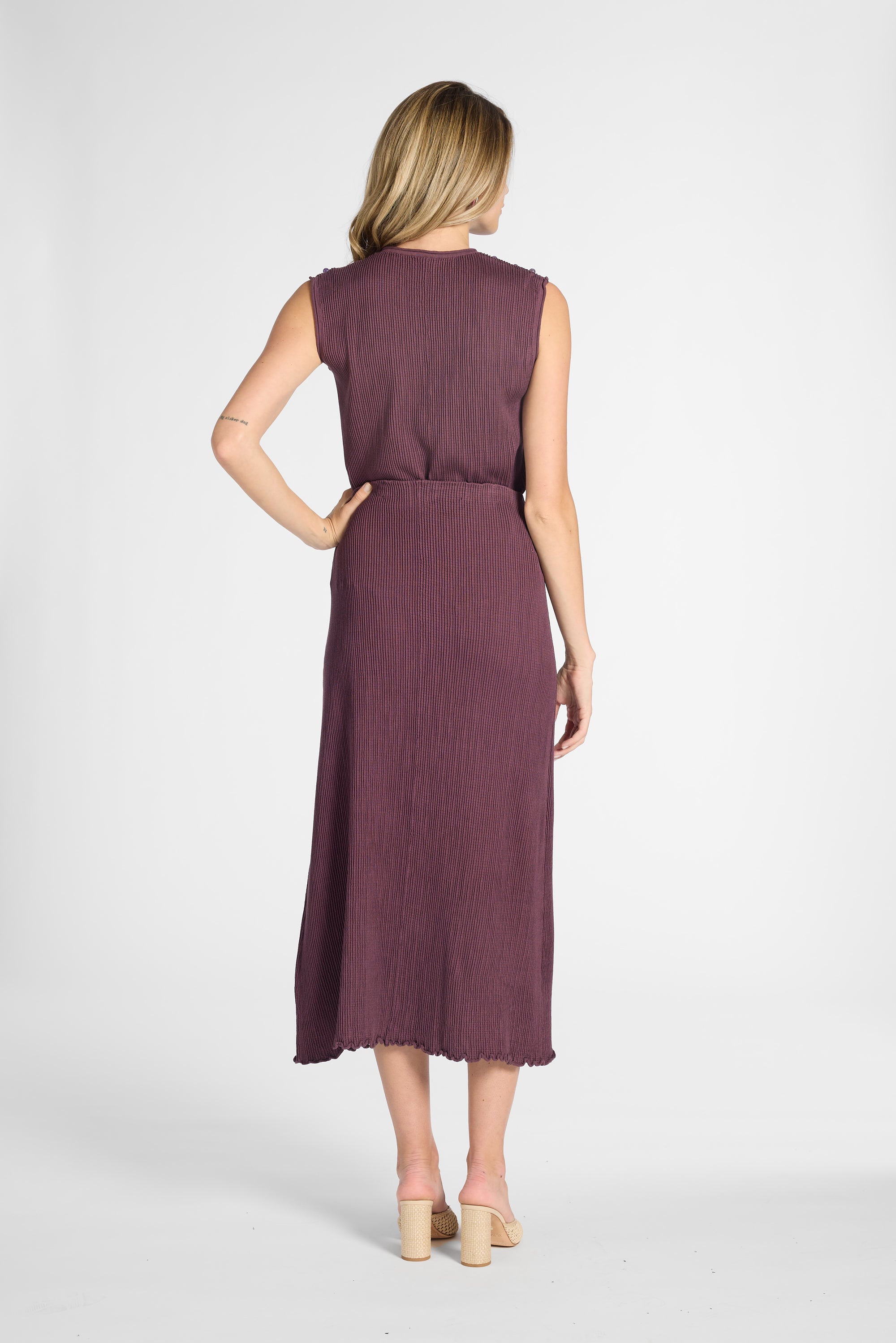 Woman standing with her back facing the camera wearing a pleated purple skirt on a white backdrop