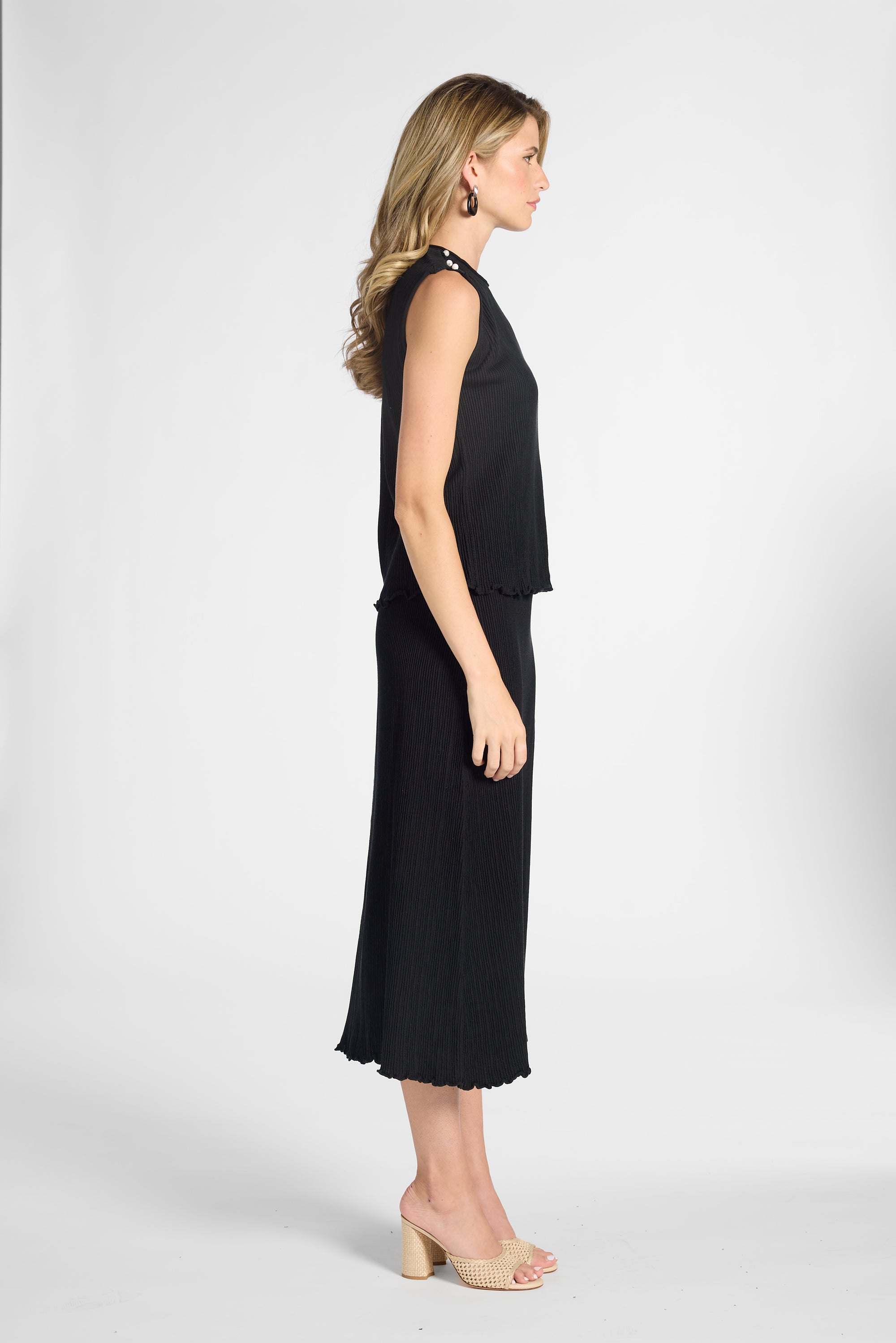 Woman standing to the side wearing a pleated black shell and the matching black skirt on a white backdrop