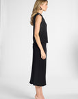 Woman standing to the side wearing a pleated black shell and the matching black skirt on a white backdrop