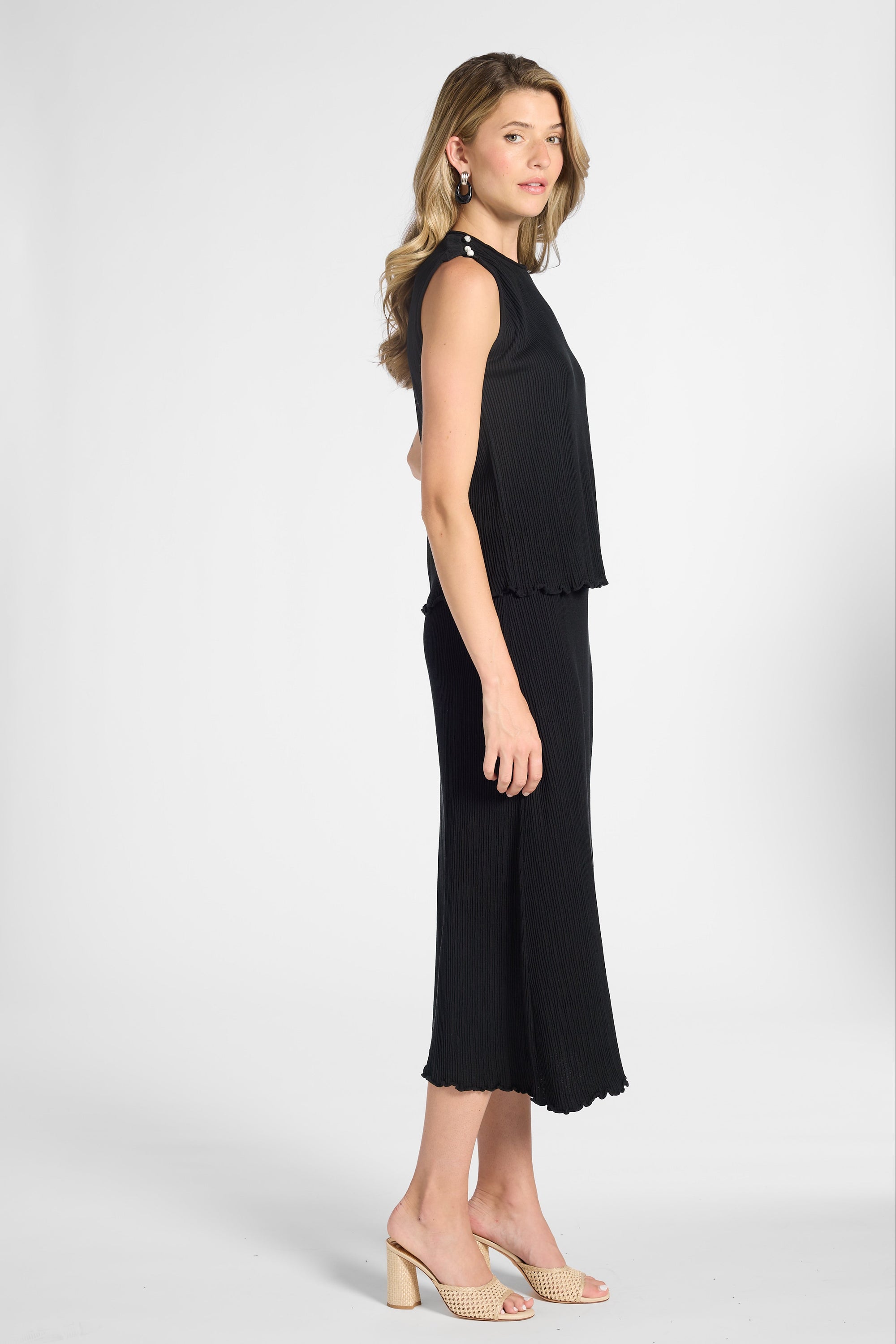 A woman modeling a sleeveless black midi dress with a ruffled peplum-style waist and a flared skirt, photographed against a white studio background.