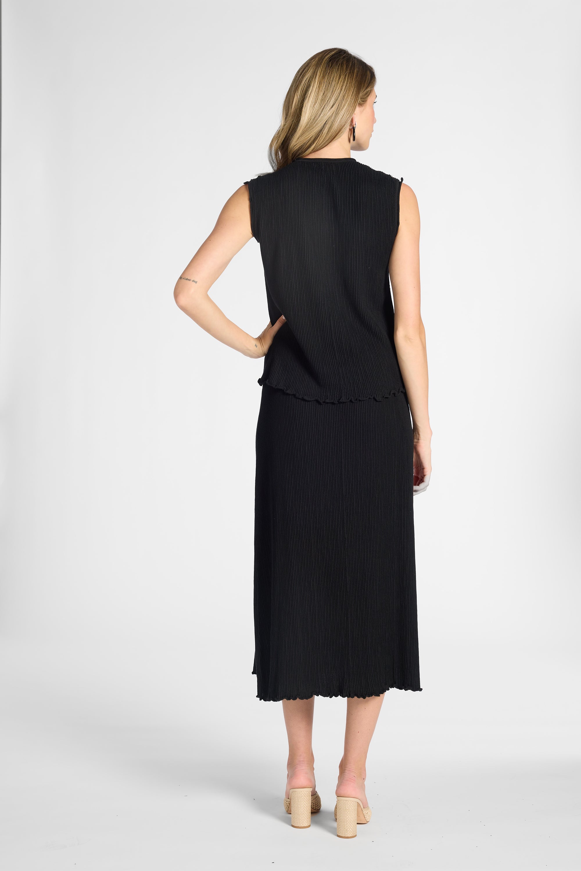 A woman shown from the back wearing a sleeveless black midi dress with a ruffled hem, paired with beige high heels, against a white studio background.
