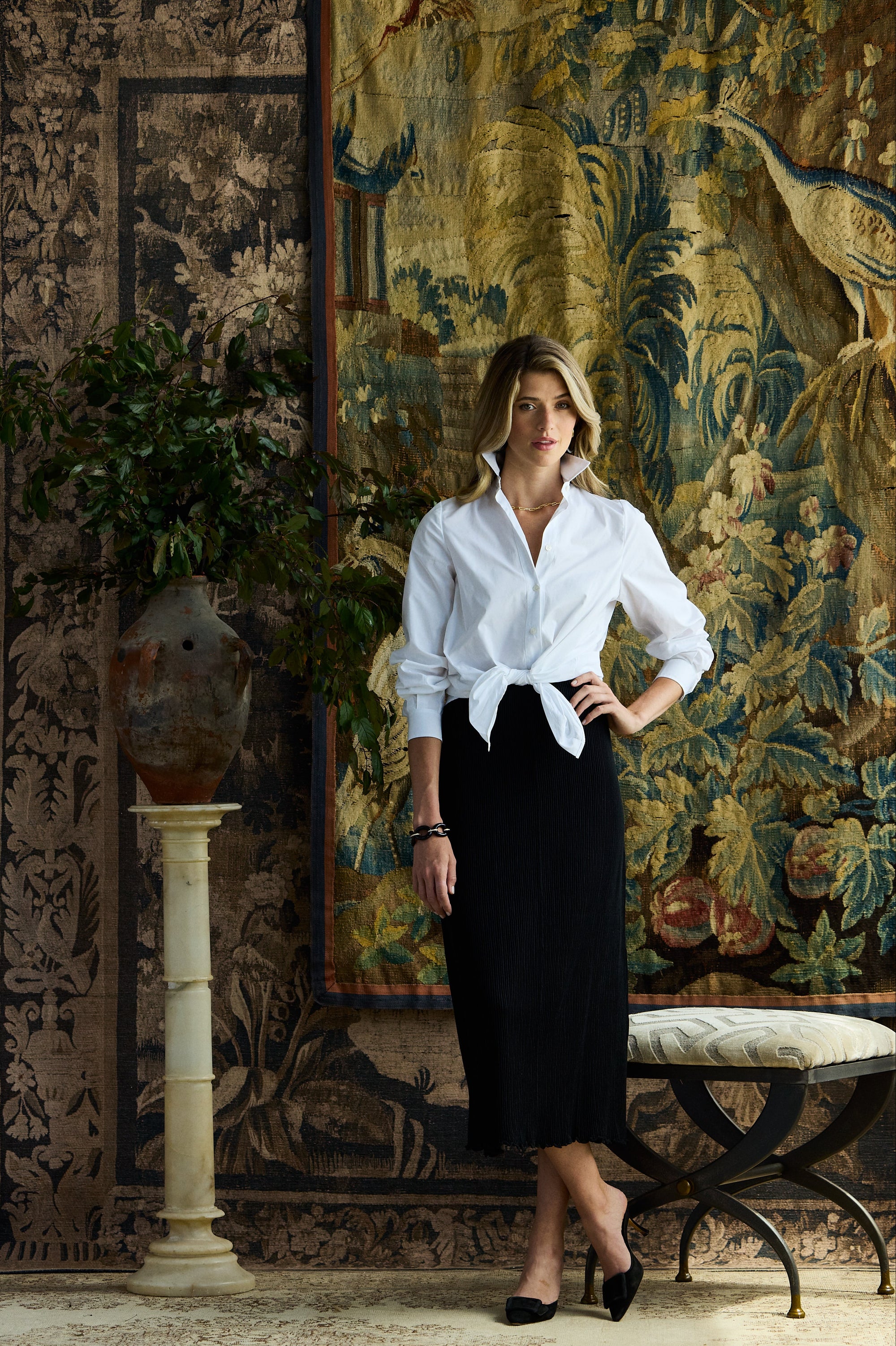 Woman standing in front of rugs wearing a black pleated skirt and a white button down blouse
