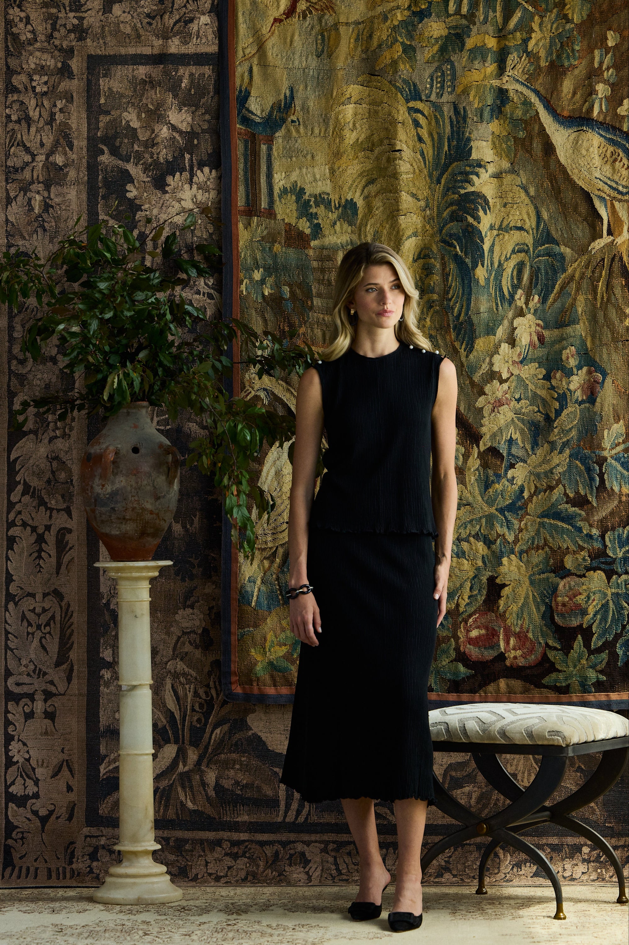 A woman stands in a styled interior wearing a sleeveless black midi dress. The background features large, ornate tapestries, a patterned rug, a potted plant on a column, and a bench.