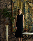 A woman stands in a styled interior wearing a sleeveless black midi dress. The background features large, ornate tapestries, a patterned rug, a potted plant on a column, and a bench.