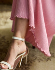A close-up of a person's foot wearing a white strappy high-heeled sandal with an ankle strap, paired with a flowing pink skirt.