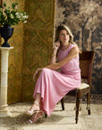 A woman wearing a sleeveless pink midi dress sits on a wooden chair in an ornate room with patterned wallpaper and a decorative screen.