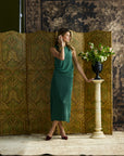 A woman models a sleeveless green midi dress while posing next to a decorative gold folding screen and a white pedestal with a flower arrangement in a vintage studio setting.