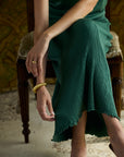 A woman seated on a wooden chair wearing a dark green ribbed knit skirt and gold jewelry, including a gold ring and bangle, in an indoor setting with patterned wallpaper.