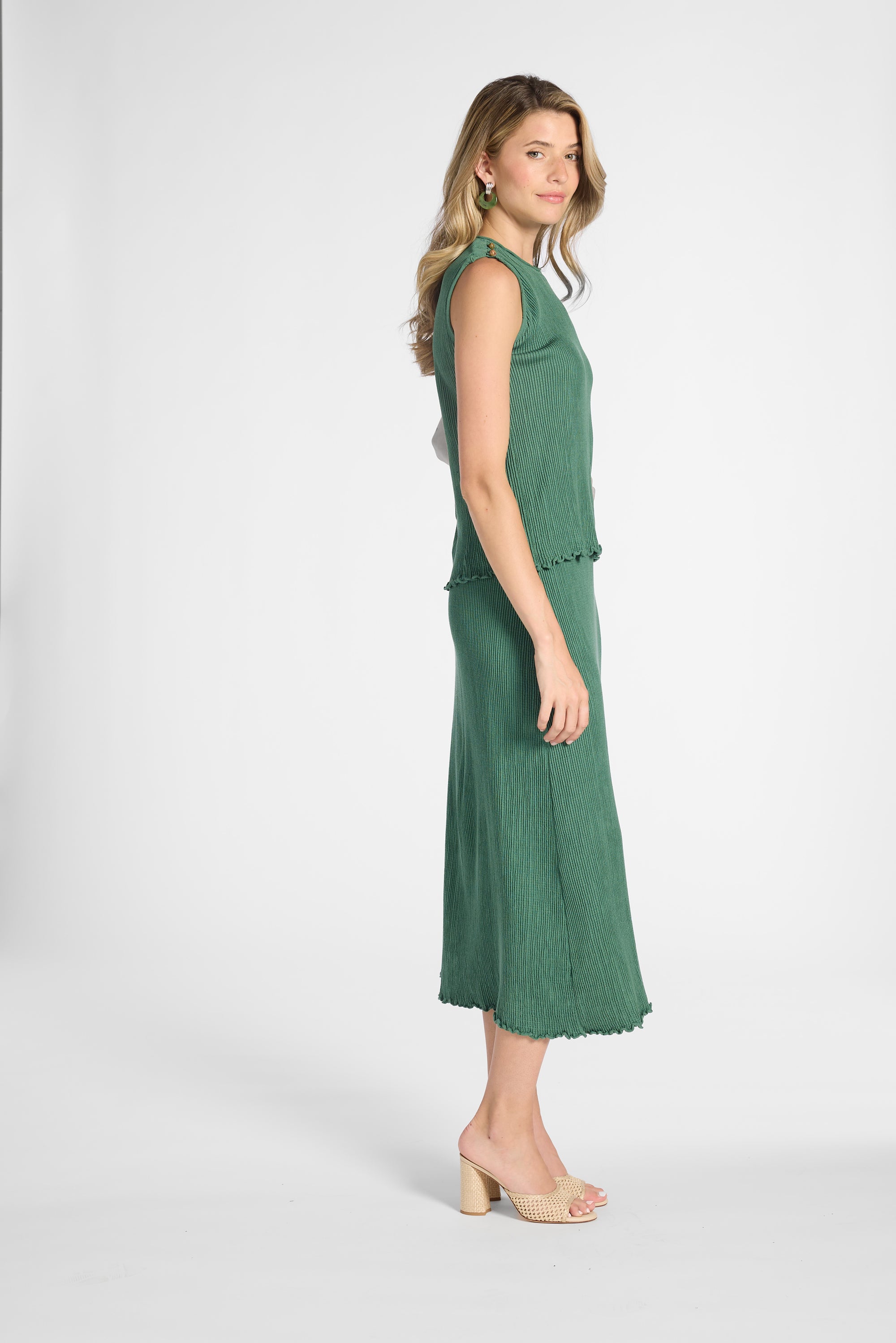 A woman modeling a sleeveless green blouse and skirt with a textured crinkle fabric and ruffled trim, shown in profile against a light gray studio background.