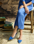 A person seated on a rustic wooden stool wearing a blue ribbed midi dress and matching pointed-toe flats with bow details, posed in an indoor setting with books and a patterned rug.