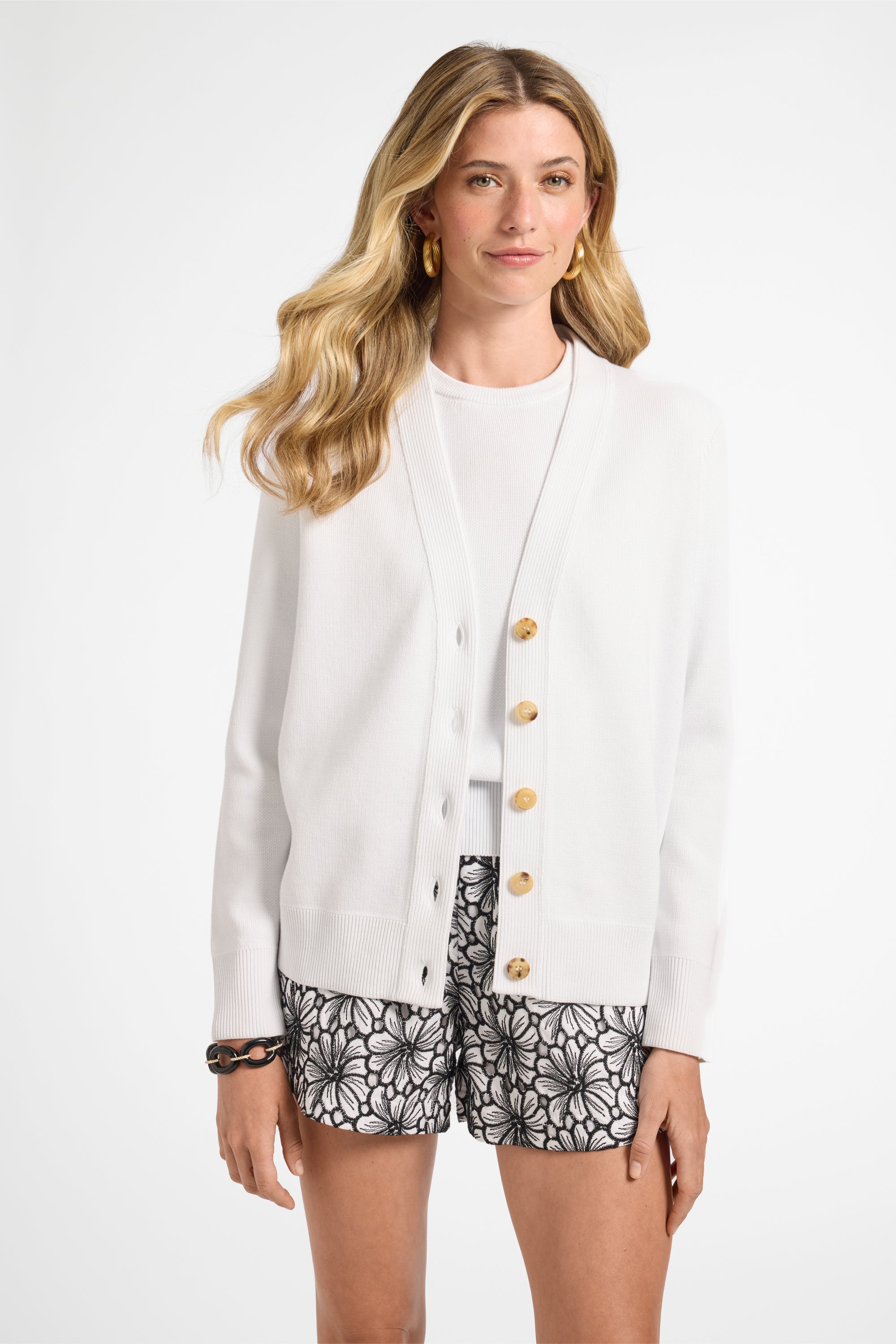 Woman wearing a white cardigan with buttons and black floral shorts on a white background