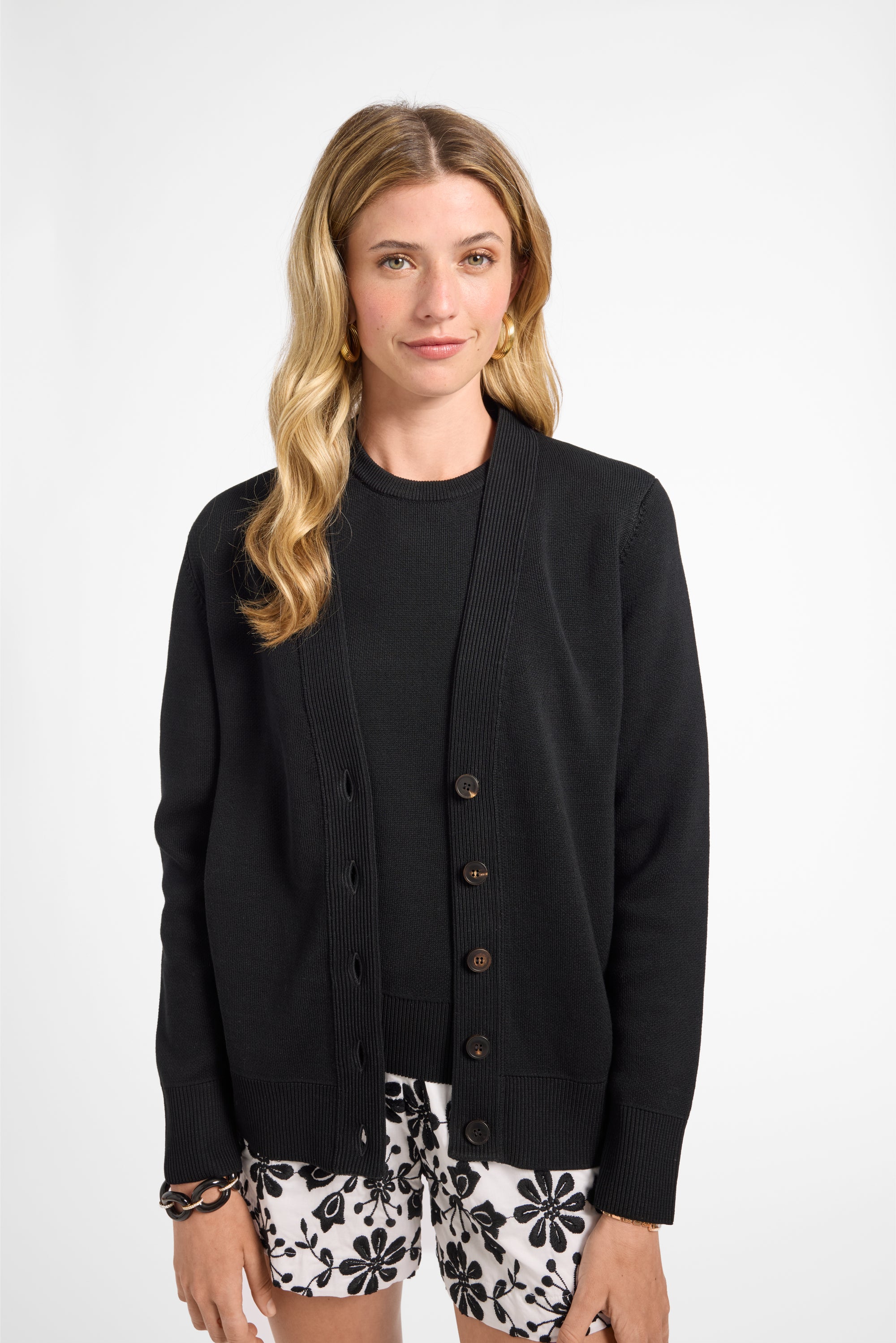 Woman wearing a black cardigan over a black top with floral shorts on a white background