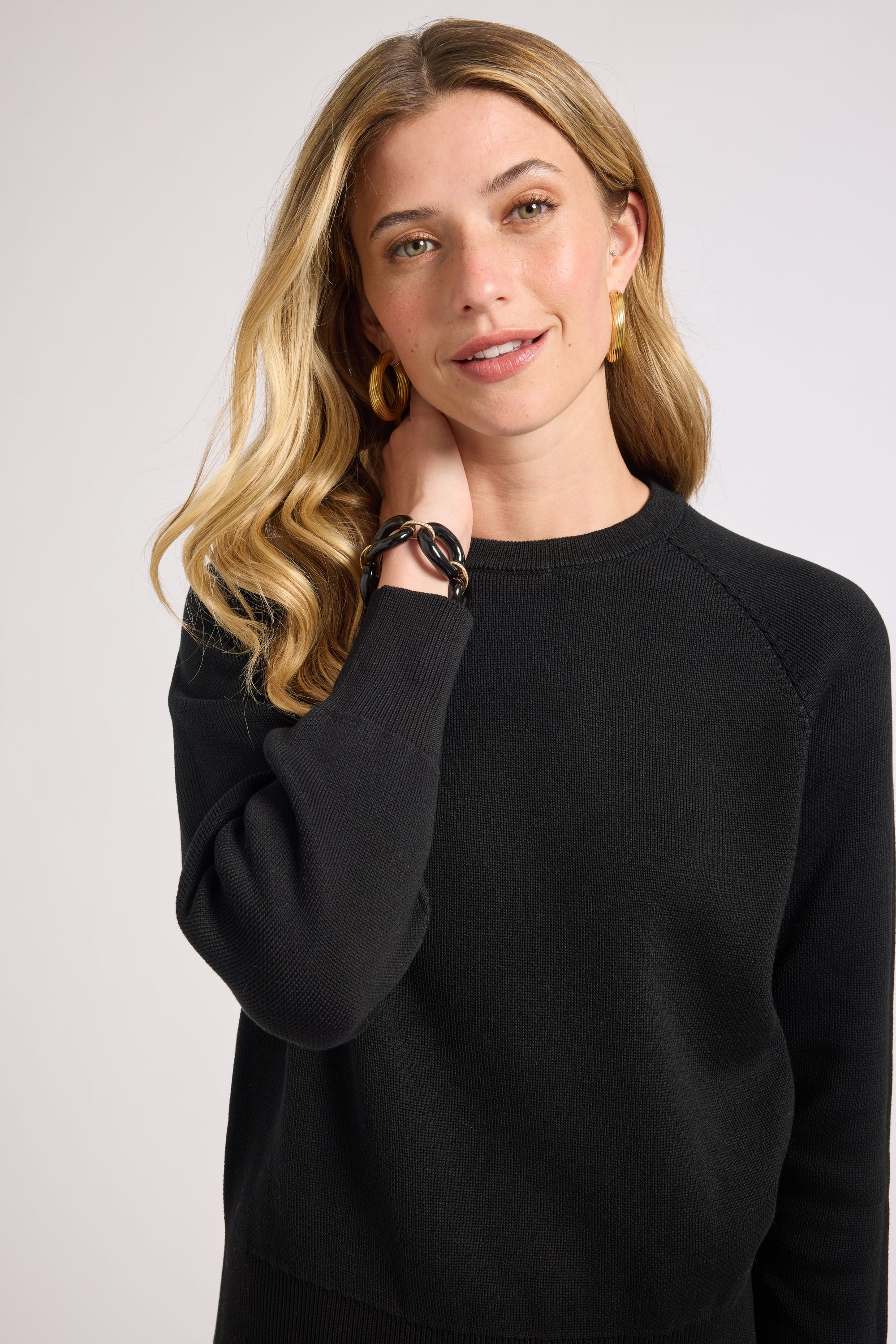Woman wearing a black sweater with gold jewelry against a plain background