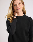 Woman wearing a black sweater with gold jewelry against a plain background