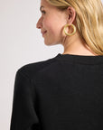 Woman wearing a black sweater and gold earrings with a plain background