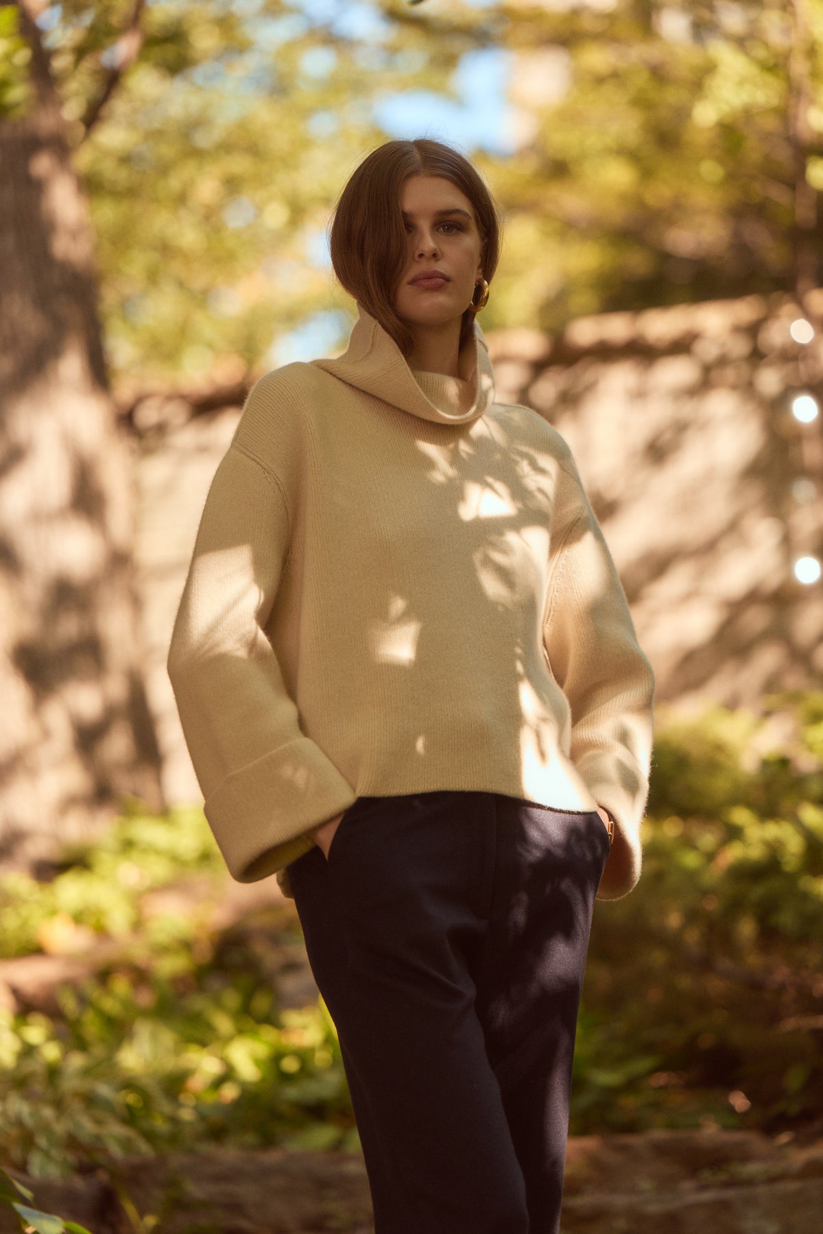 A woman wearing a cream-colored cowl-neck knit sweater and dark navy trousers, standing outdoors in a sun-dappled wooded setting.