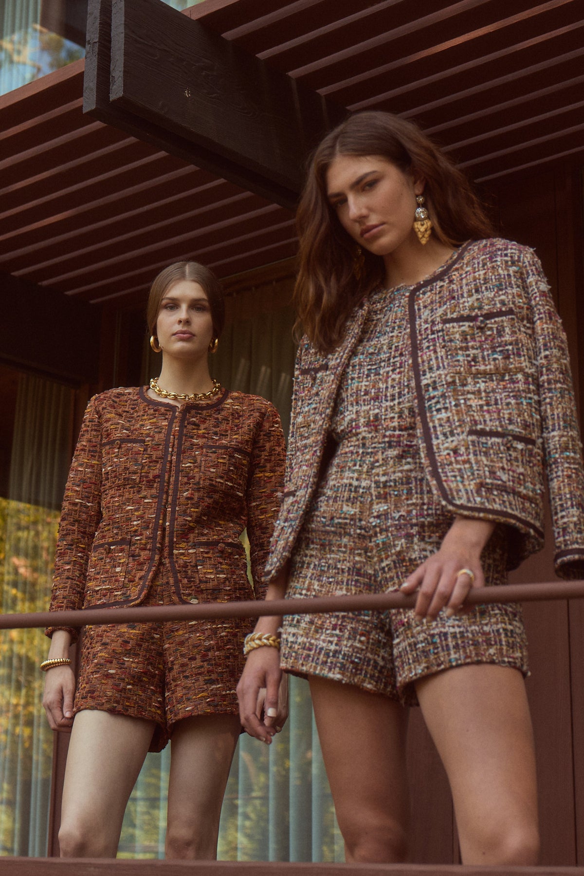 Two women modeling matching tweed mini dresses and shorts sets on a modern wooden balcony with large windows and trees visible outside.