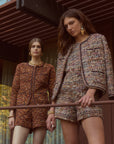 Two women modeling matching tweed mini dresses and shorts sets on a modern wooden balcony with large windows and trees visible outside.