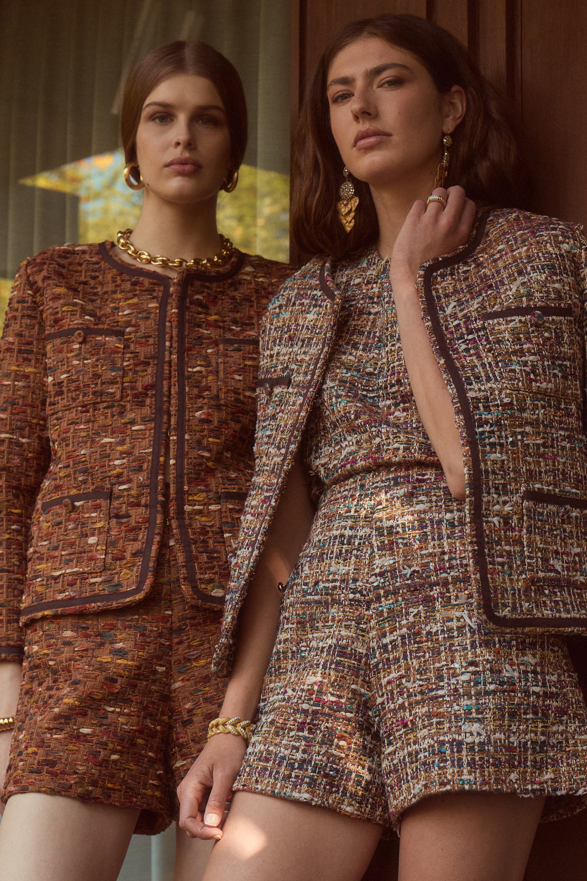Two women modeling matching tweed mini suits in an outdoor setting with warm-toned glass doors and trees visible in the background. The suits feature a textured multicolor tweed pattern with brown, beige, and gold tones.