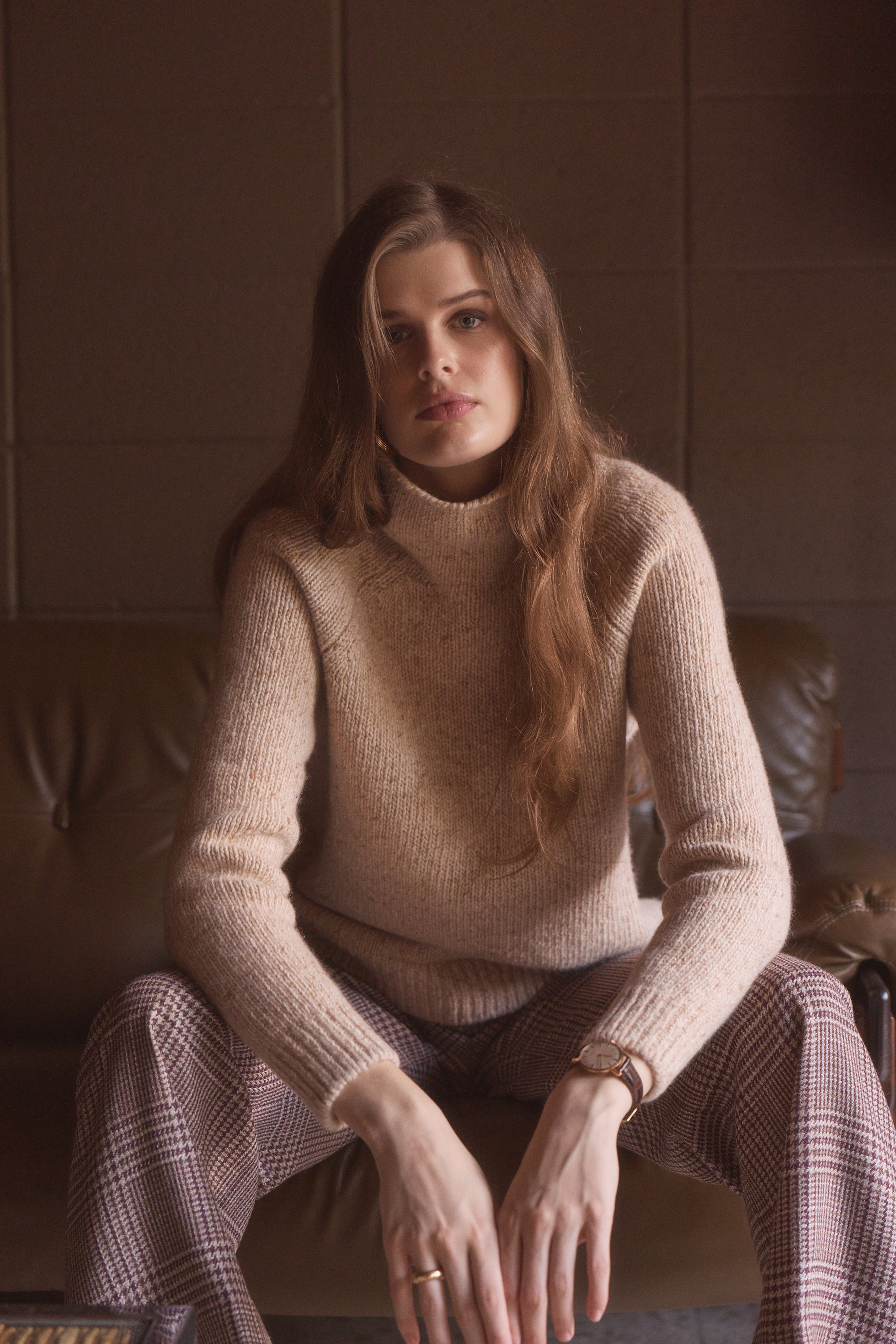 Woman modeling a tan cashmere turtleneck sweater while sitting on a couch