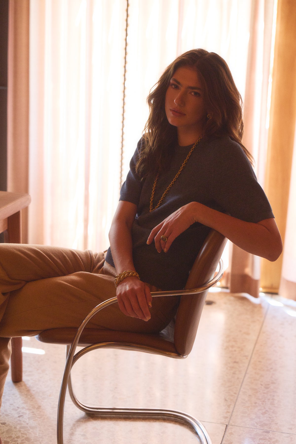 Woman in a house sitting in a chair modeling a blue cotton tee with brown pants
