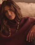 A woman with long wavy brown hair reclines on a beige couch, wearing a chunky burgundy turtleneck sweater and a gold chain necklace.