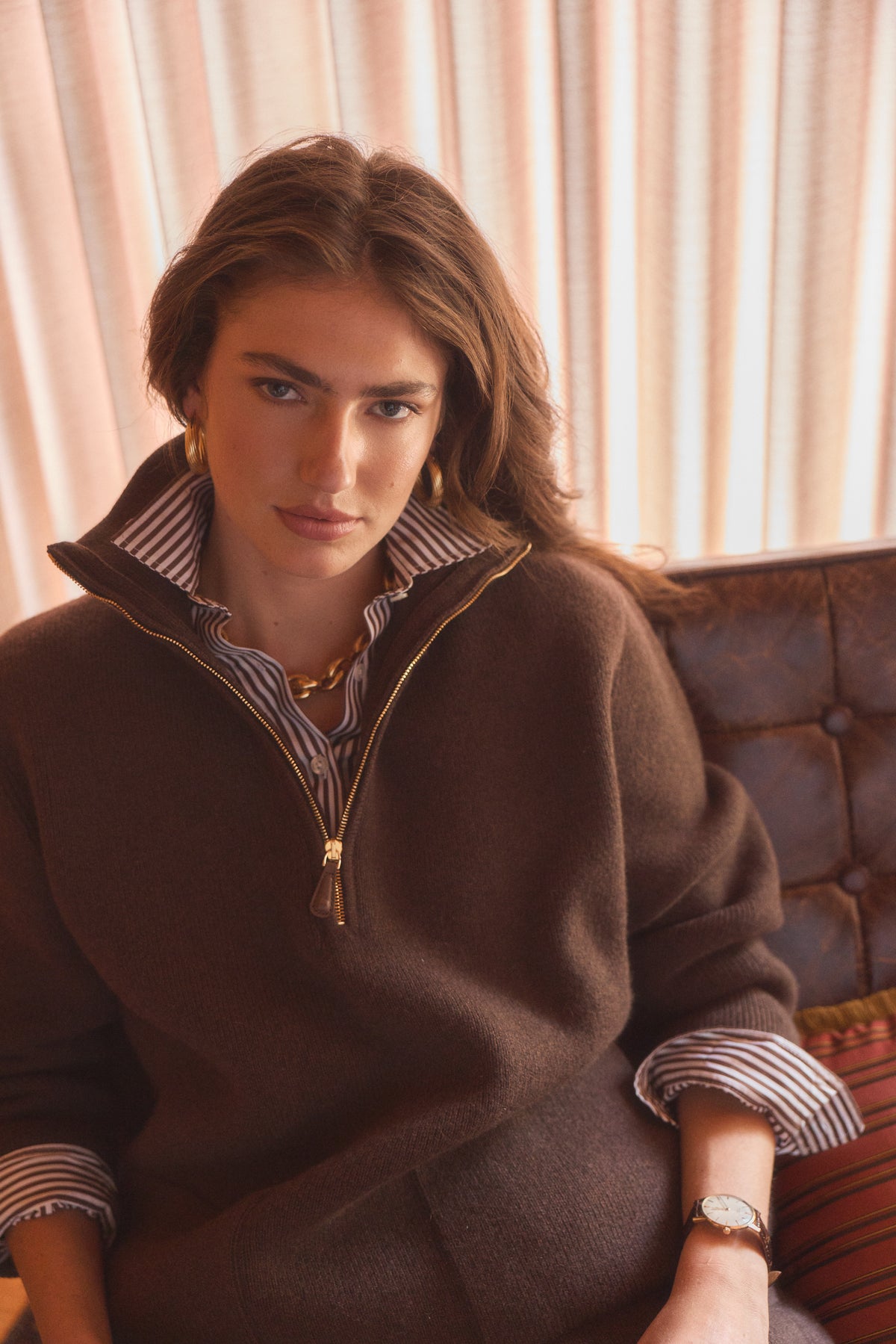 A woman wearing a brown quarter-zip sweater over a striped collared shirt, seated on a brown leather sofa against a beige curtain background.