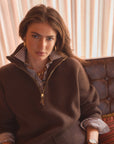A woman wearing a brown quarter-zip sweater over a striped collared shirt, seated on a brown leather sofa against a beige curtain background.