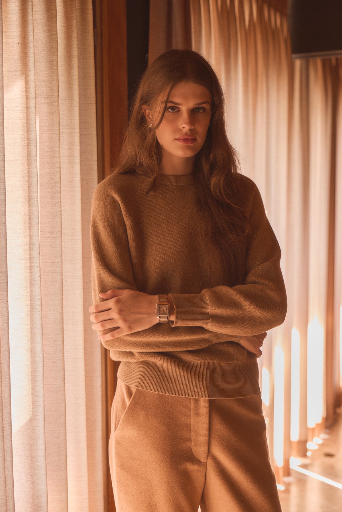Woman modeling a tan crewneck in a home in front of curtains