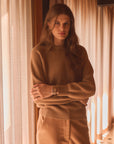 Woman modeling a tan crewneck in a home in front of curtains