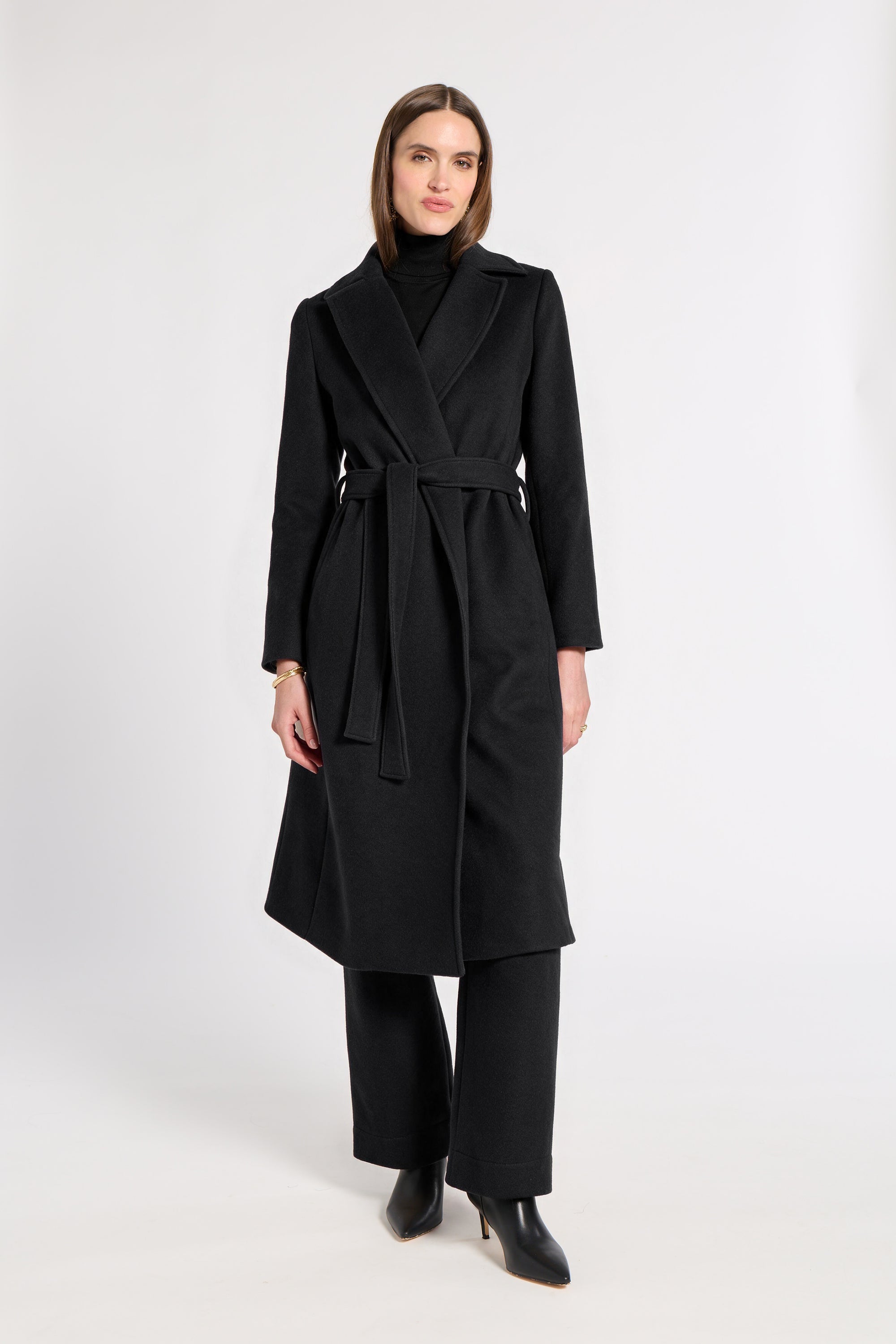 A woman modeling a long black wrap-style cashmere coat with a matching waist tie, worn over a black turtleneck and black wide-leg trousers, against a plain light gray studio background.