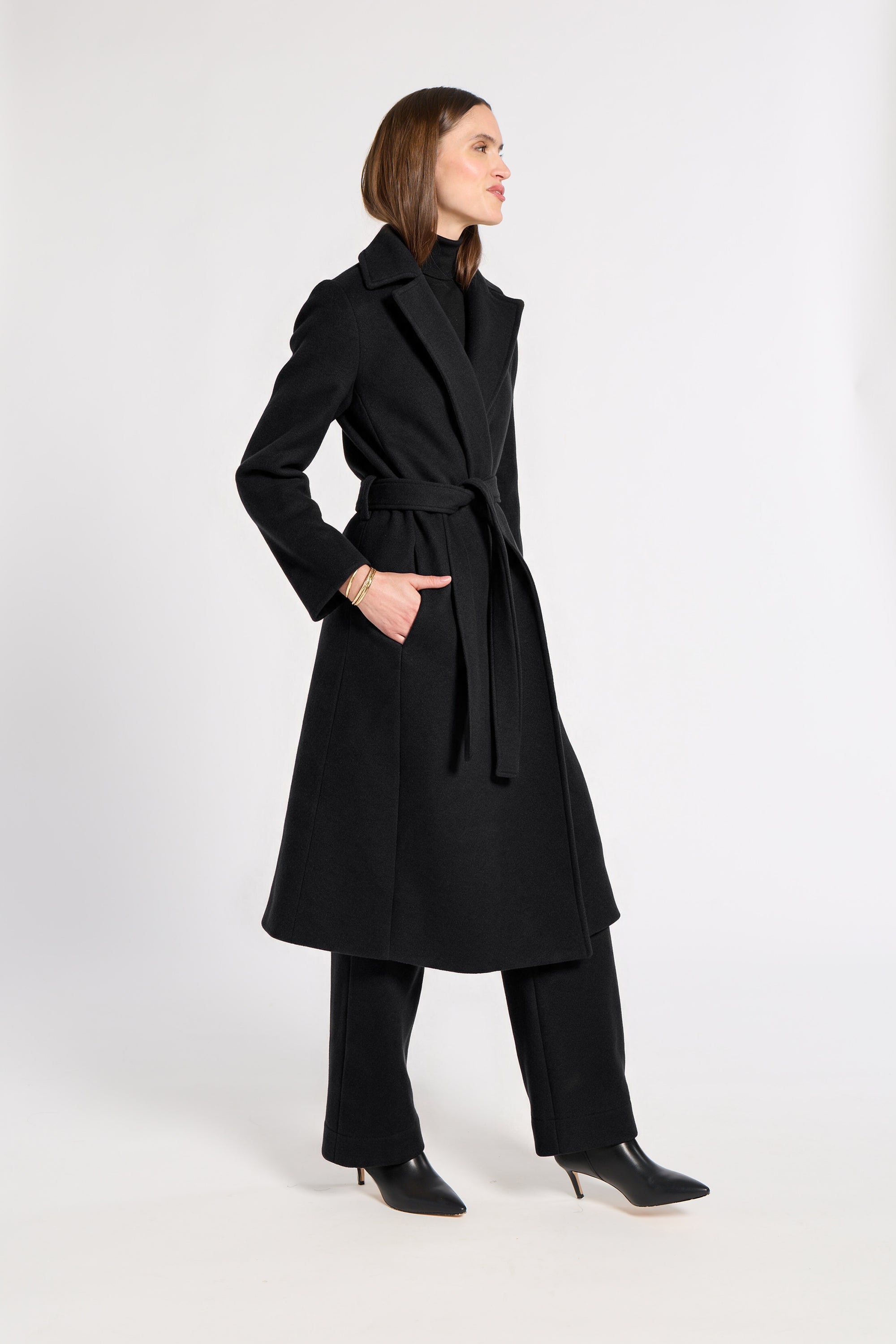 A woman modeling a long black belted coat, black wide-leg trousers, and black pointed-toe heels against a plain light gray studio background.