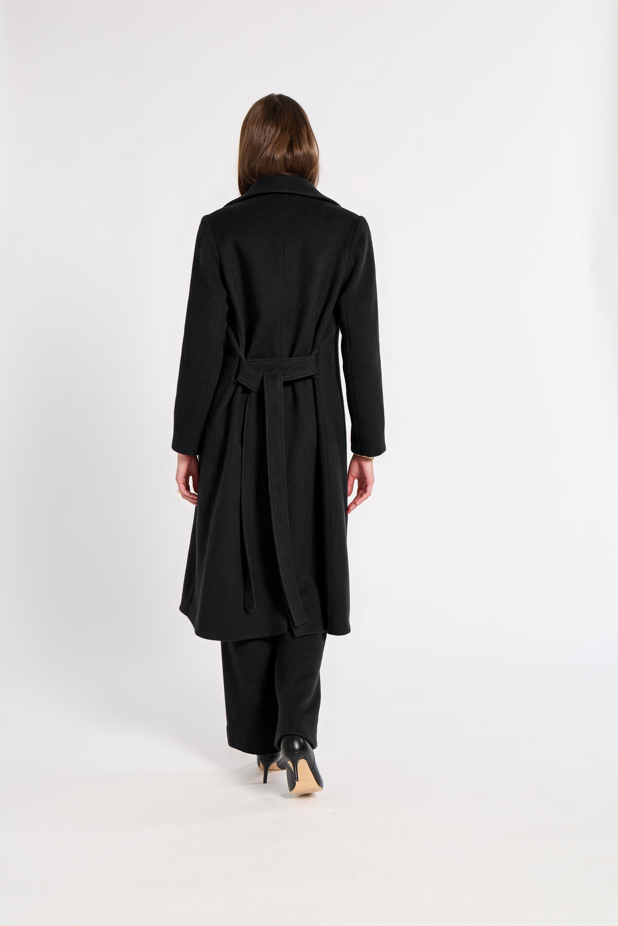 A woman viewed from behind wearing a long black belted coat, black pants, and black high-heeled shoes against a plain white studio background.
