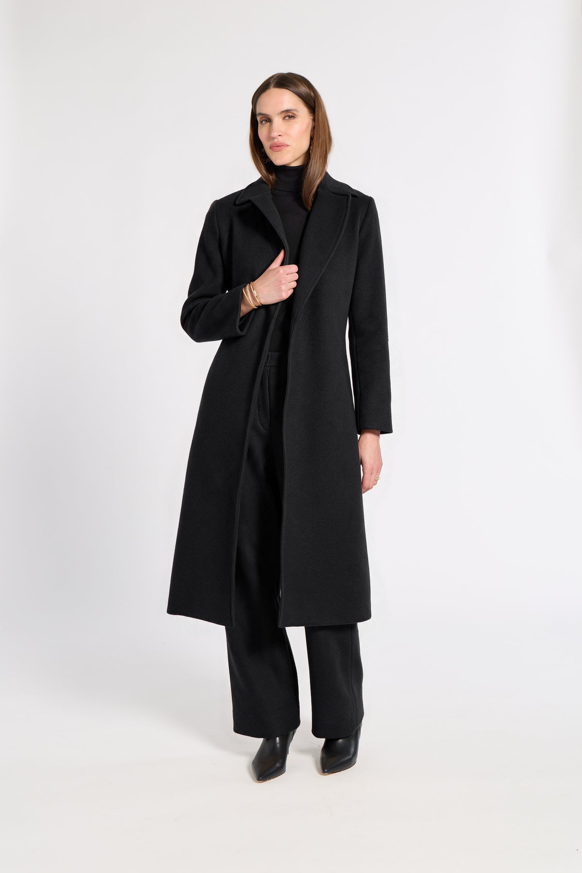 A woman modeling a long black coat over a black turtleneck and wide-leg black trousers, standing against a plain light gray studio background.