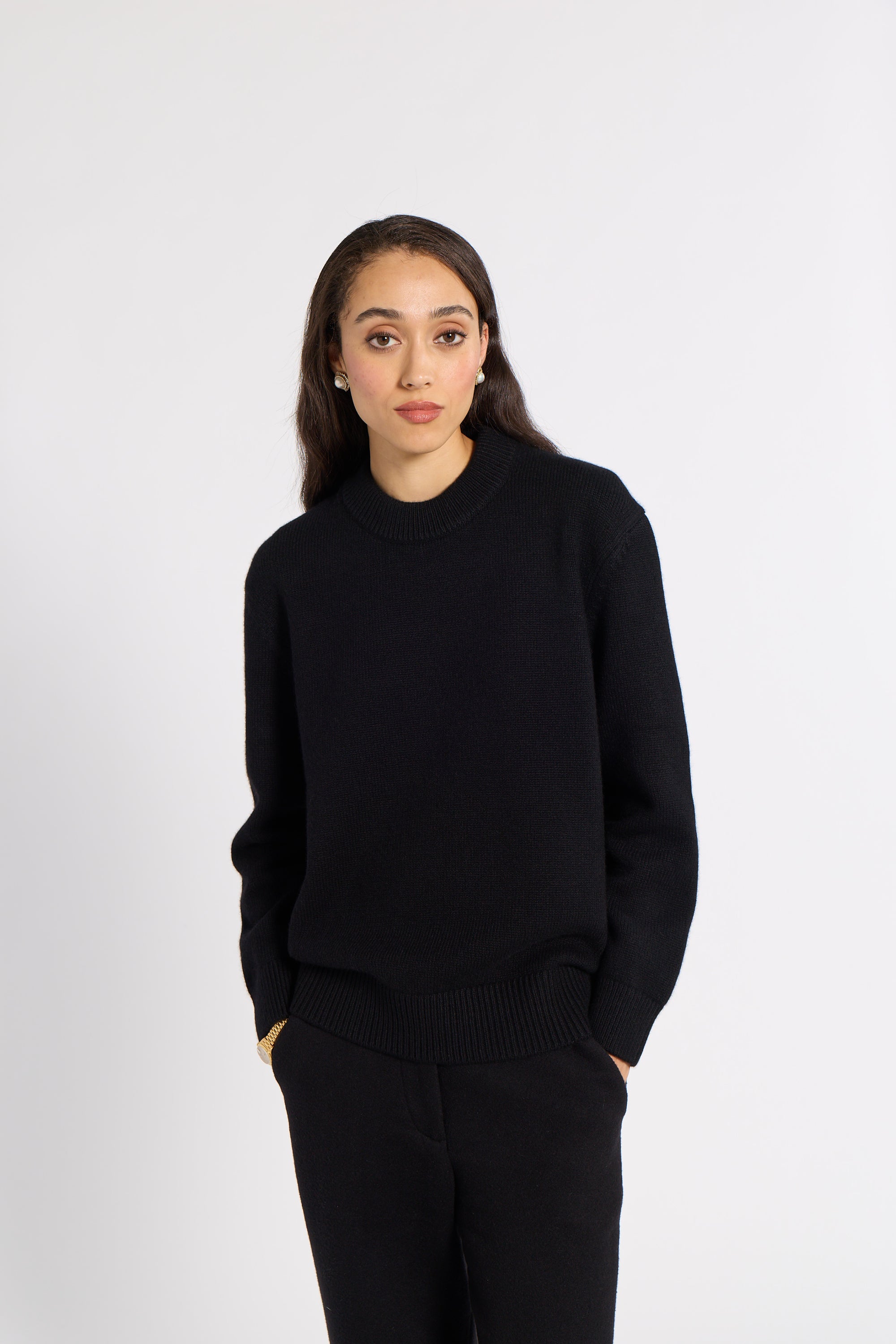 A woman modeling a black crewneck knit sweater and matching black trousers against a plain light gray studio background.
