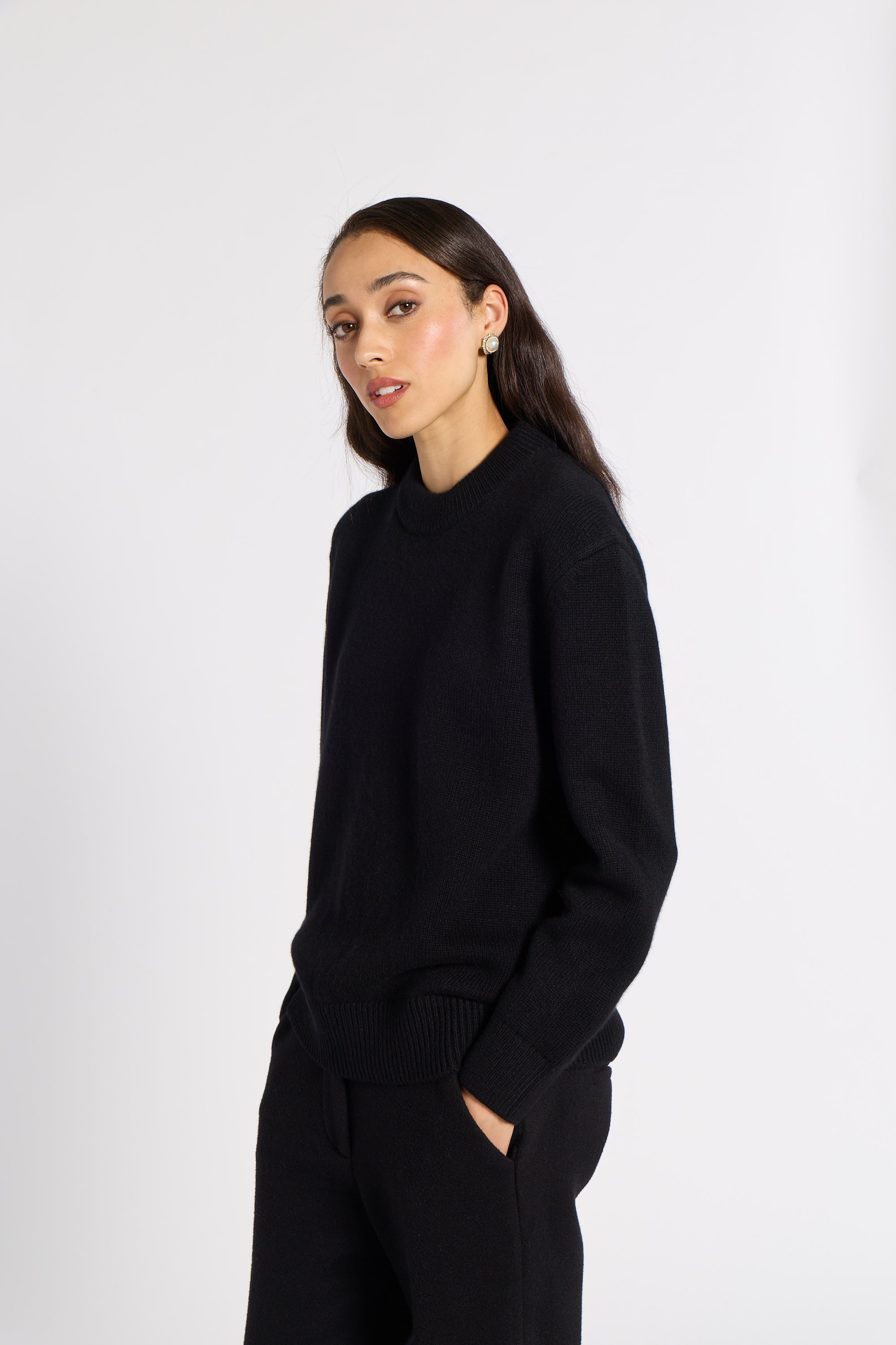 A woman modeling a black knit mock neck sweater and matching black knit pants against a plain light gray studio background.
