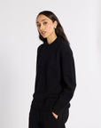 A woman modeling a black knit mock neck sweater and matching black knit pants against a plain light gray studio background.