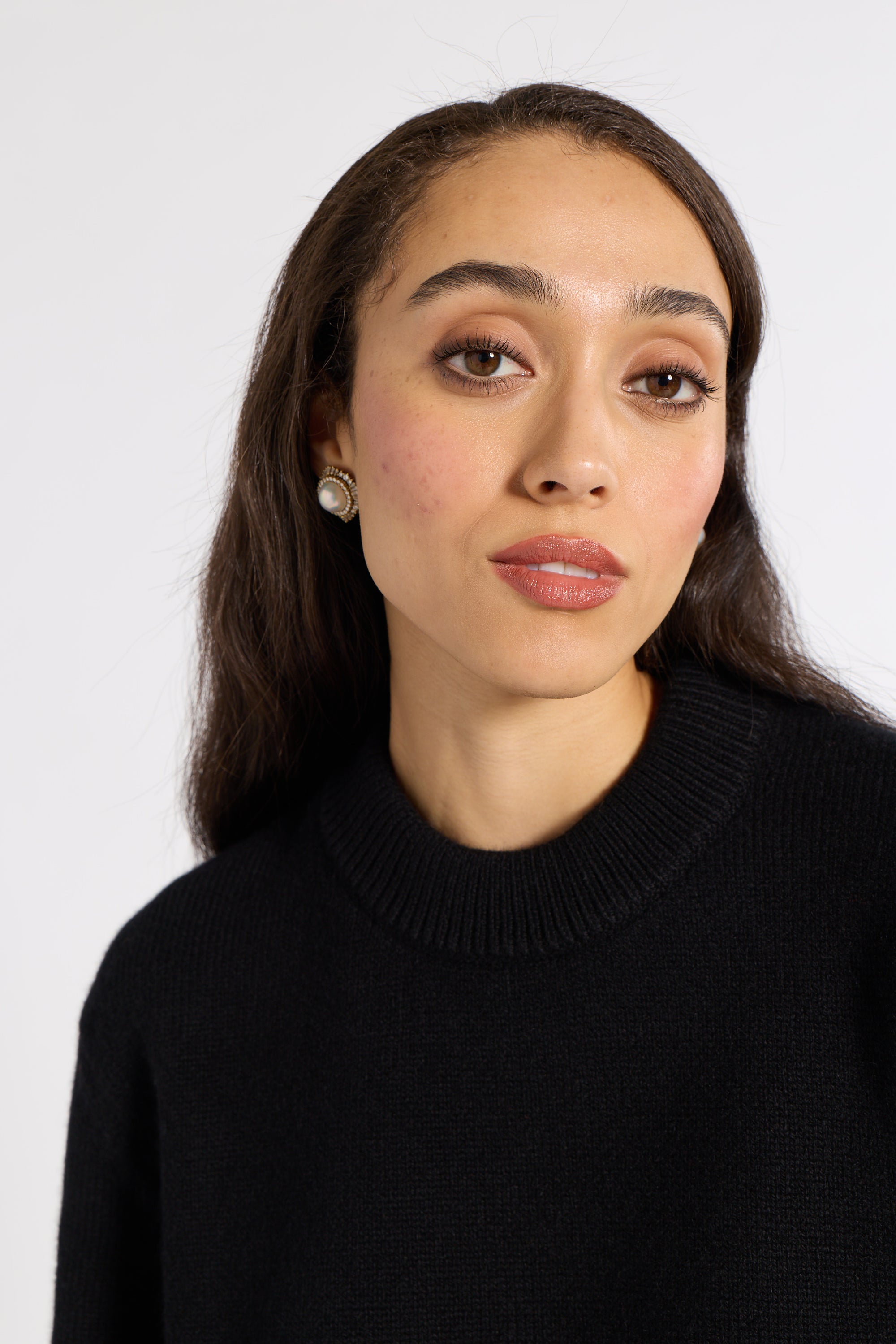 A close-up portrait of a woman with dark hair wearing a black ribbed knit mock neck sweater against a plain light gray background.