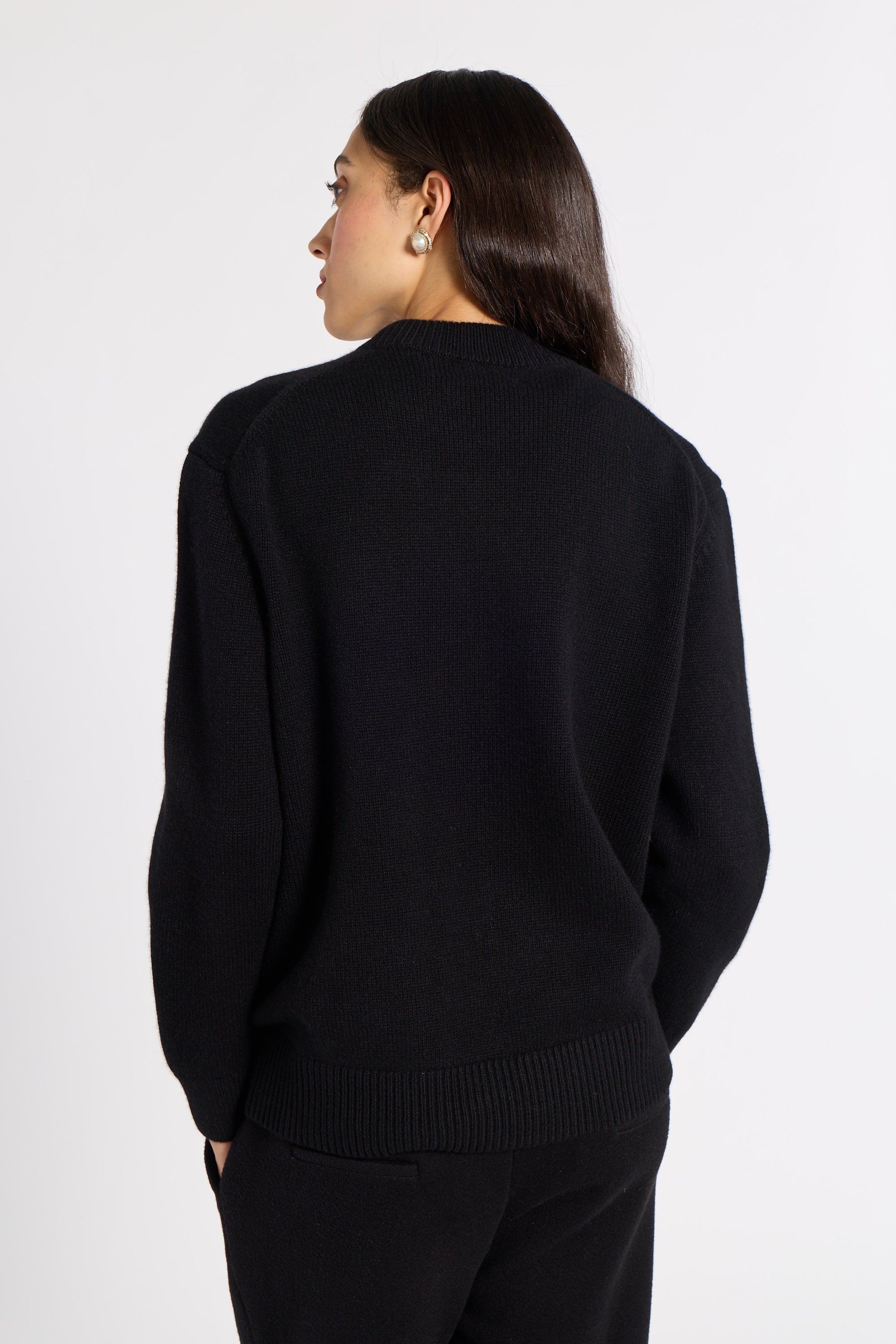 A woman shown from the back wearing a black knit sweater and black pants against a plain light gray studio background.
