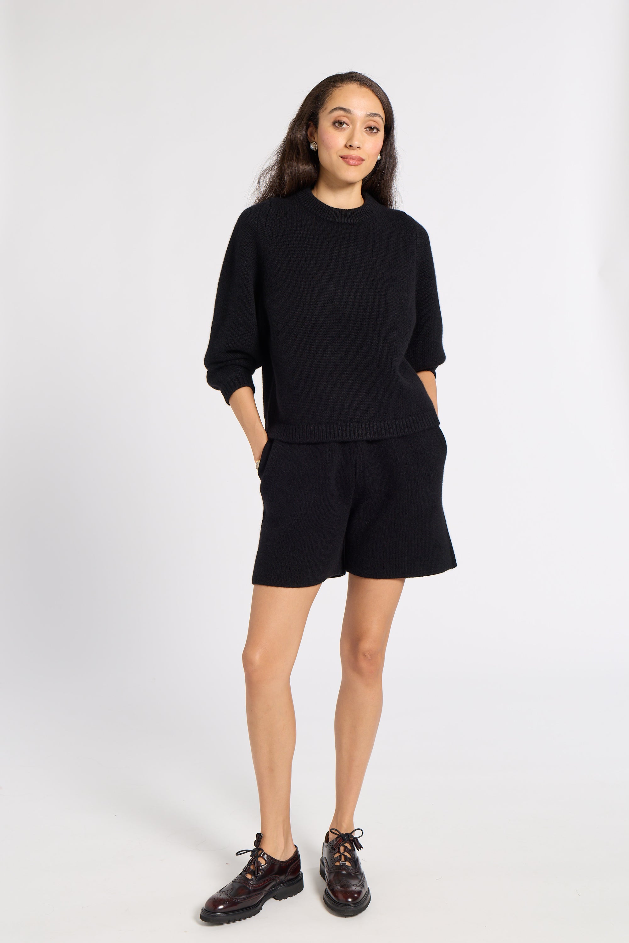 A woman modeling a black knit two-piece lounge set consisting of a crewneck sweatshirt and matching shorts, paired with dark brown lace-up shoes, against a light gray studio background.