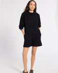 A woman modeling a black knit two-piece lounge set consisting of a crewneck sweatshirt and matching shorts, paired with dark brown lace-up shoes, against a light gray studio background.