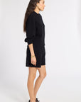 A woman stands in profile wearing a black knit sweater and matching black shorts, paired with dark brown leather shoes, against a plain light gray studio background.