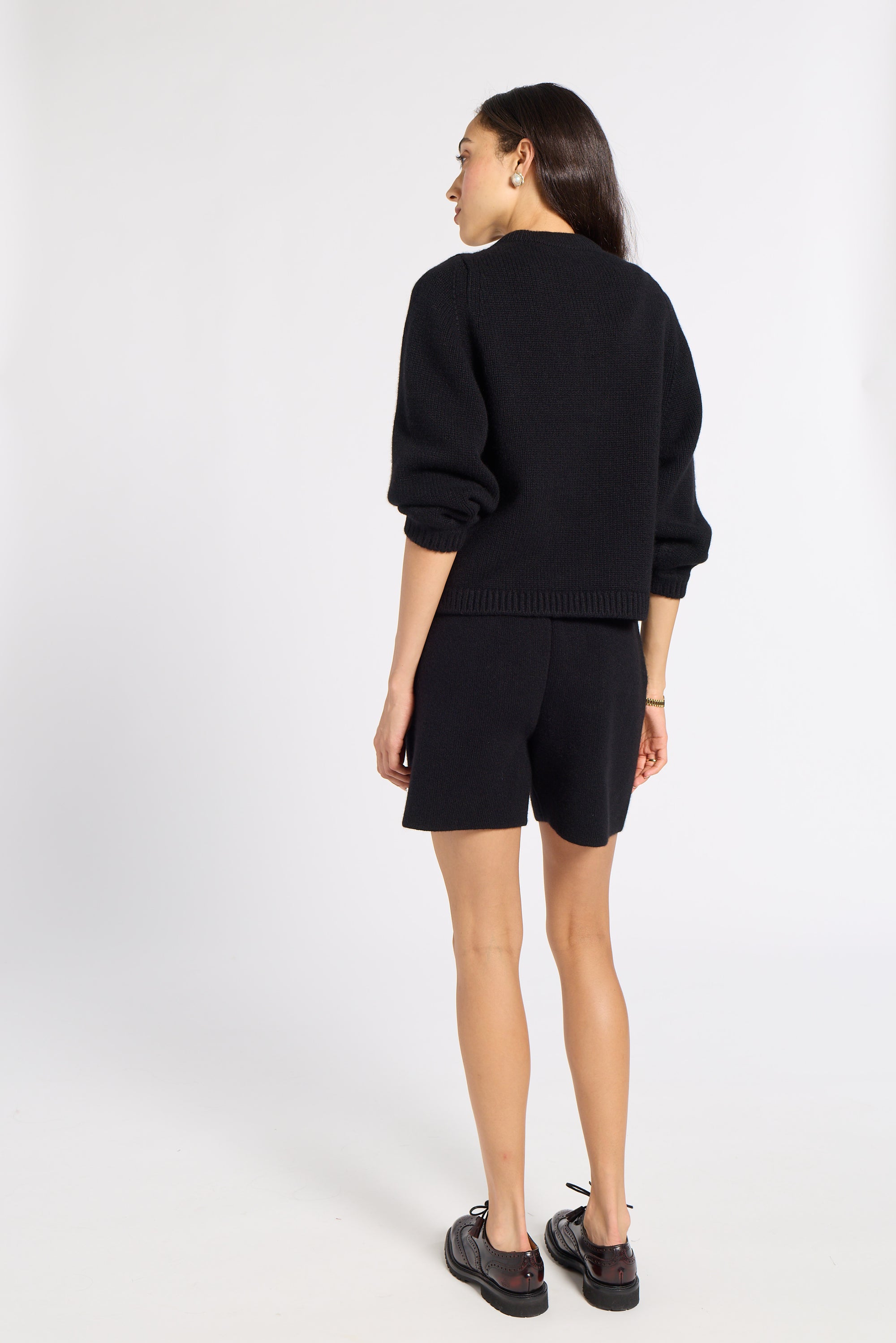 A woman is shown from the back wearing a black knit sweater and matching black shorts, paired with dark brown shoes, against a plain white studio background.