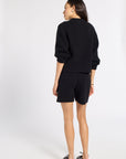 A woman is shown from the back wearing a black knit sweater and matching black shorts, paired with dark brown shoes, against a plain white studio background.