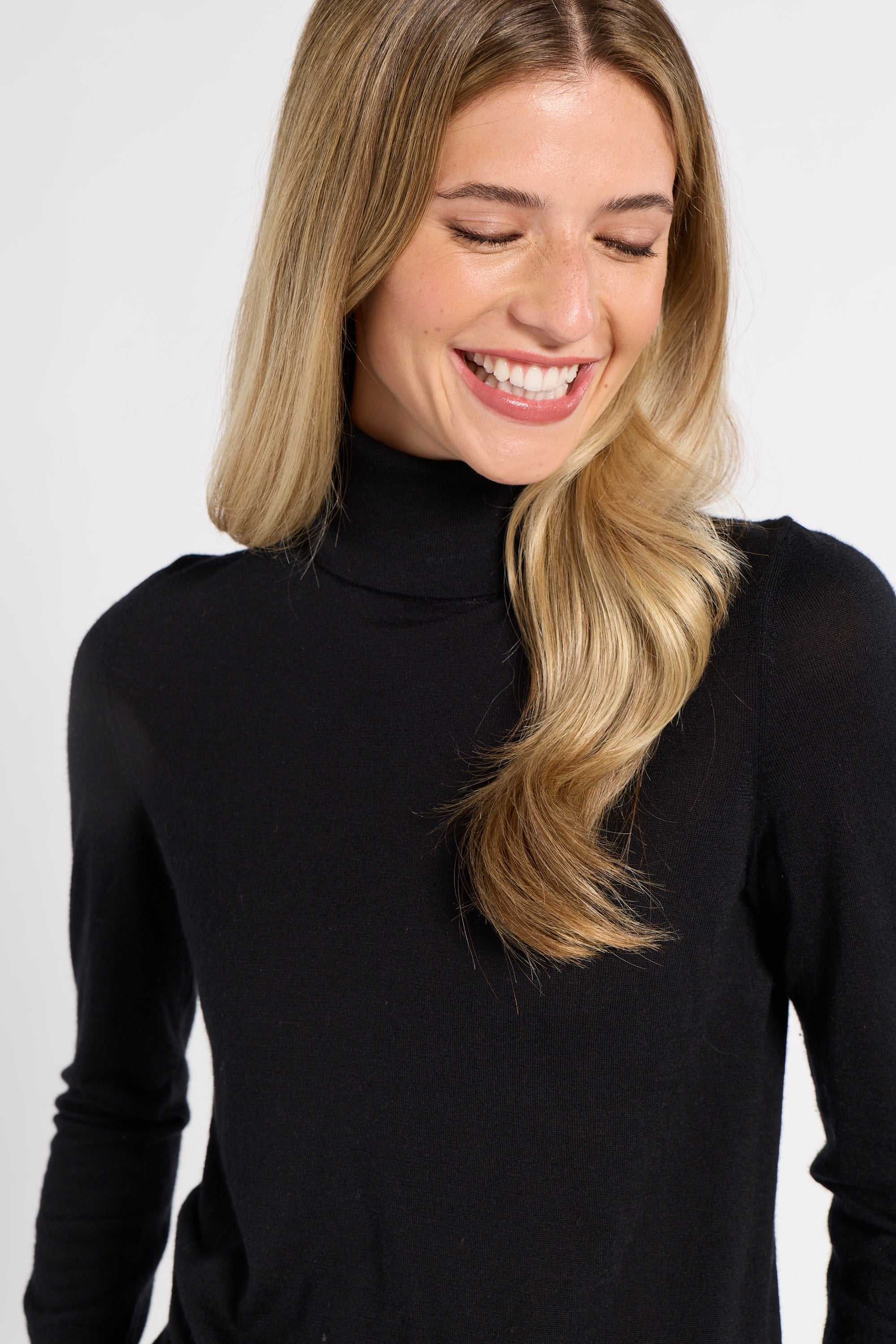 A smiling blonde woman wearing a black long-sleeve mock neck turtleneck sweater, shown from the chest up against a light gray studio background.
