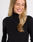 A smiling blonde woman wearing a black long-sleeve mock neck turtleneck sweater, shown from the chest up against a light gray studio background.