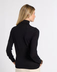A woman with blonde hair is shown from the back wearing a black long-sleeve mock neck top and cream pants against a plain white studio background.