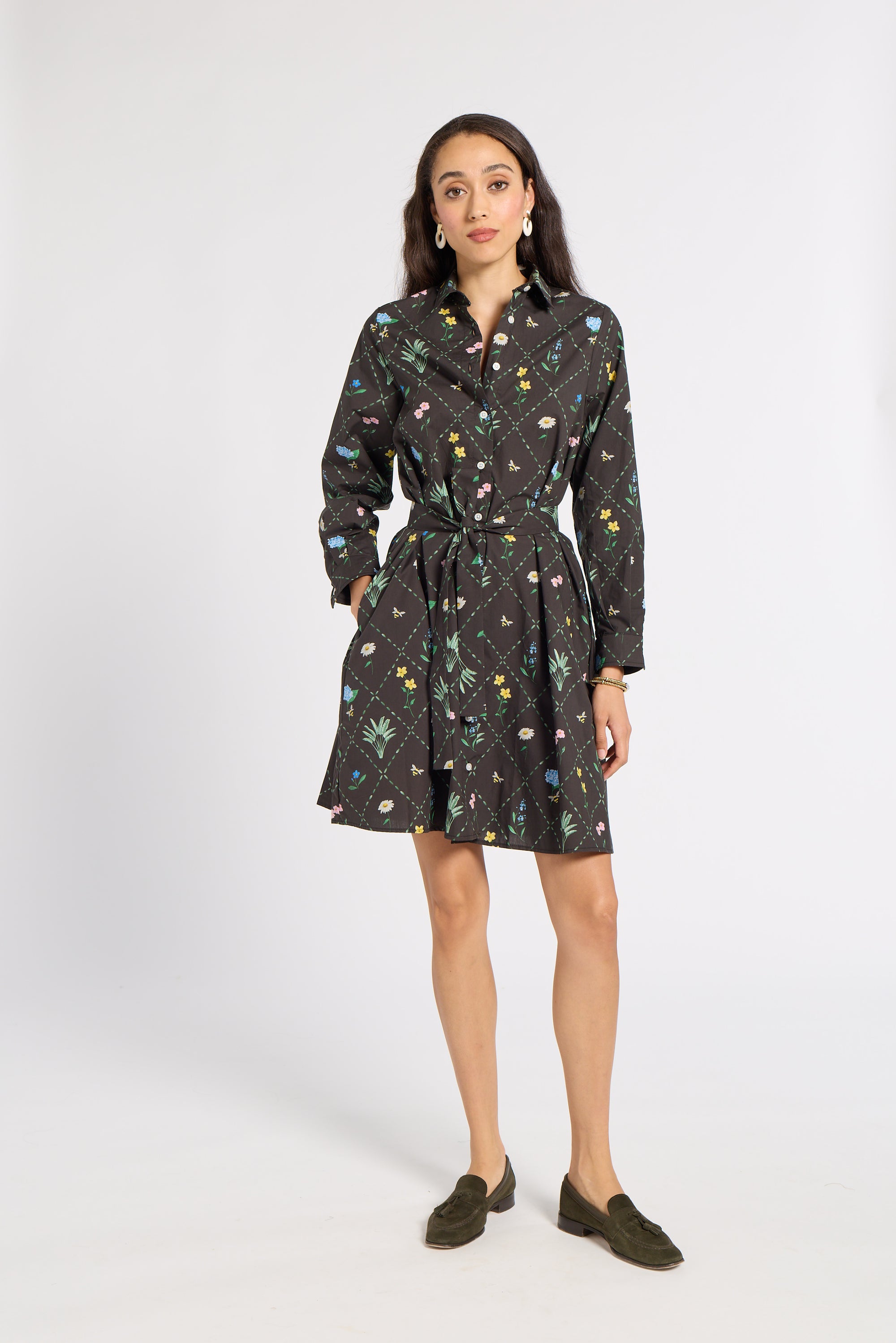A woman modeling a black long-sleeve mini dress with a colorful floral and botanical print, featuring a tie waist belt and collar, paired with dark olive green loafers against a light gray studio background.